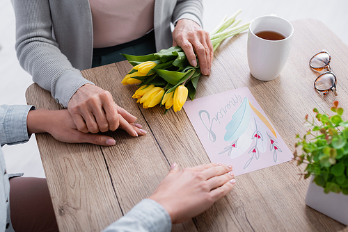 Cropped view of senior woman holding hand of daughter and flowers near tea and greeting card with 8 march lettering