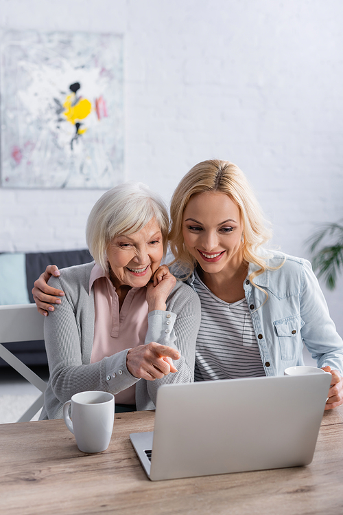Senior woman pointing with finger near daughter with cup and laptop