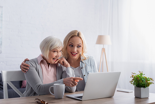 Cheerful elderly woman pointing at laptop near cup and daughter