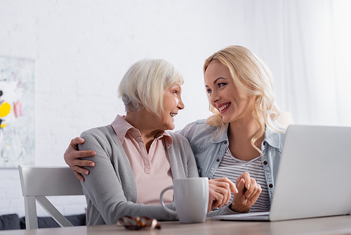 Cheerful senior woman looking at daughter near laptop and cup