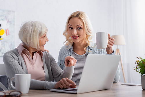 Smiling woman holding cup near mother pointing at laptop at home