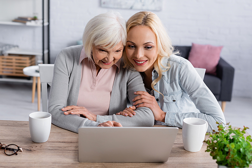 Smiling woman and mother using laptop near cups on table