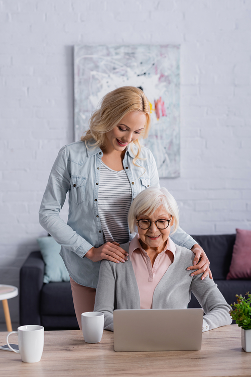 Cheerful woman hugging senior mother in eyeglasses using laptop near cups