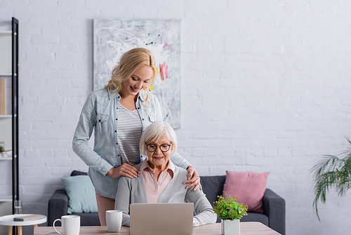 Smiling woman standing near senior mother using laptop in living room