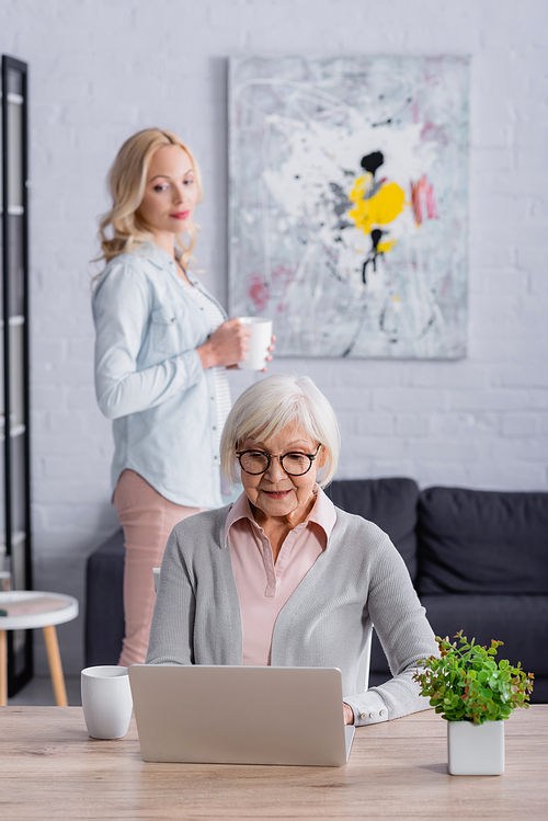 Senior woman using laptop near cup and daughter on blurred background at home