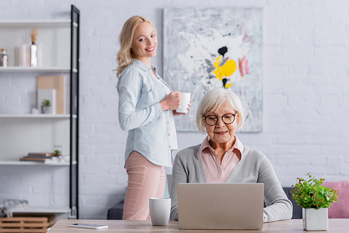 Positive grey haired woman using laptop near daughter with tea on blurred background