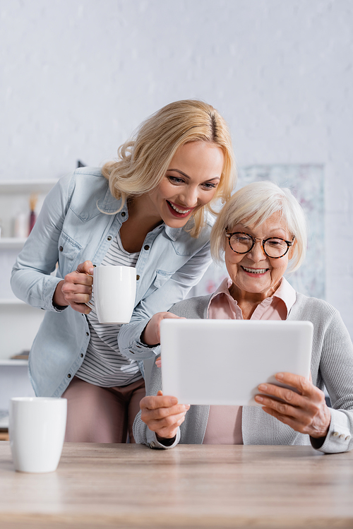 Smiling woman with cup standing near senior parent using digital tablet