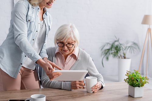 Positive woman pointing at digital tablet near elderly mom with cup at home
