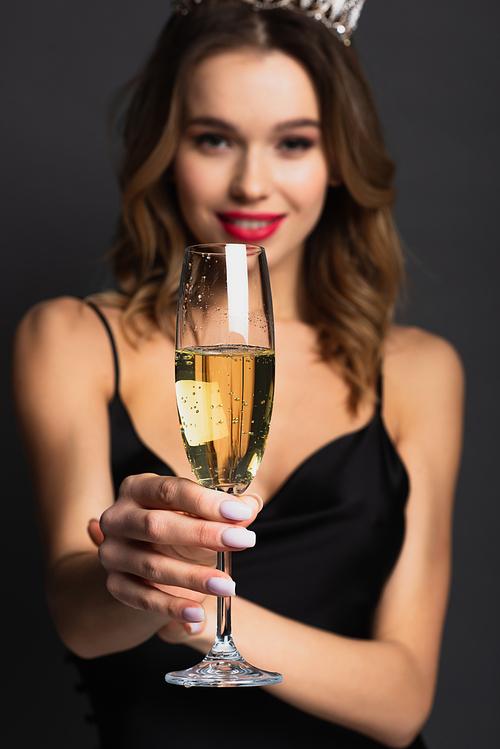 blurred happy woman in black slip dress and tiara holding glass of champagne on grey