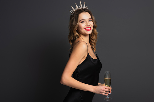 happy young woman in black slip dress and tiara holding glass of champagne on grey