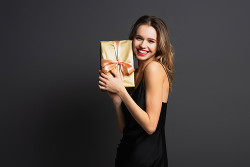 happy young woman in black slip dress holding wrapped gift box on grey