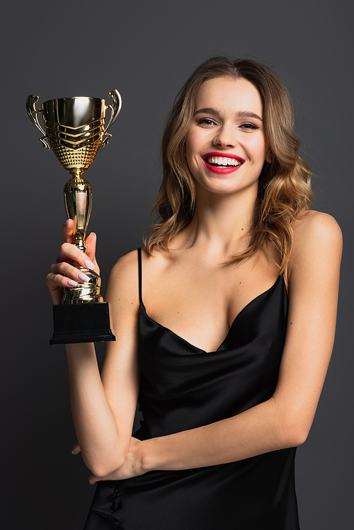 happy young woman in black slip dress holding golden trophy on grey