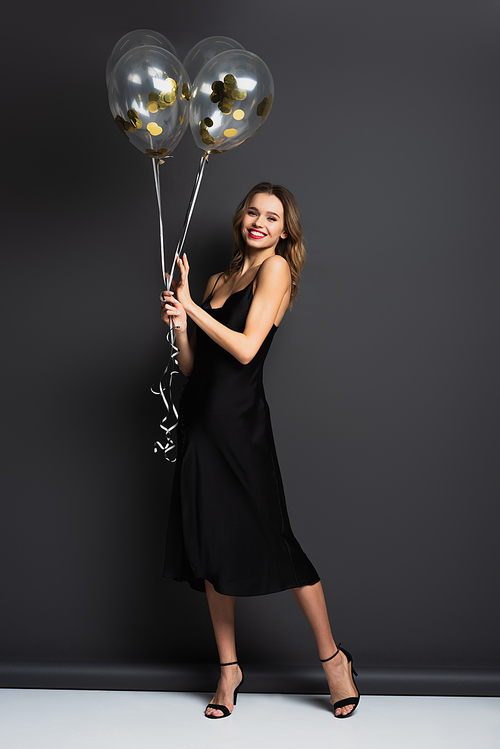 full length of smiling young woman in black slip dress holding balloons on grey
