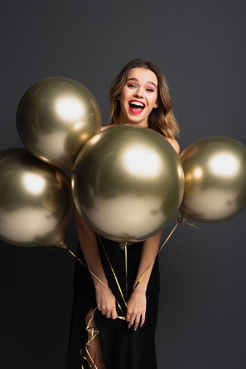 happy young woman in black slip dress holding golden balloons on grey