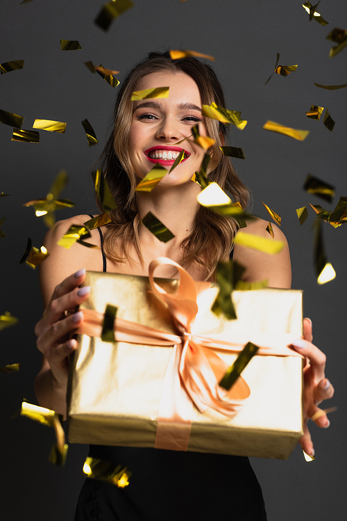 joyful young woman in black slip dress holding gift box near blurred confetti on grey