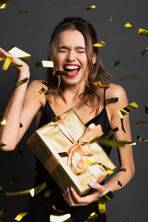excited young woman in black slip dress holding gift box near blurred confetti on grey