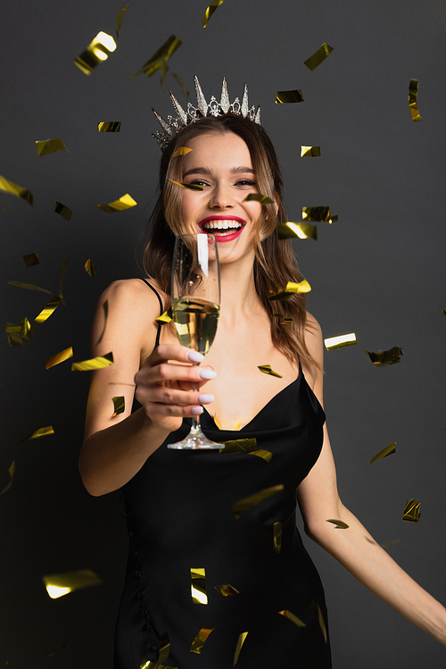 joyful woman in slip dress and tiara holding glass of champagne near confetti on grey