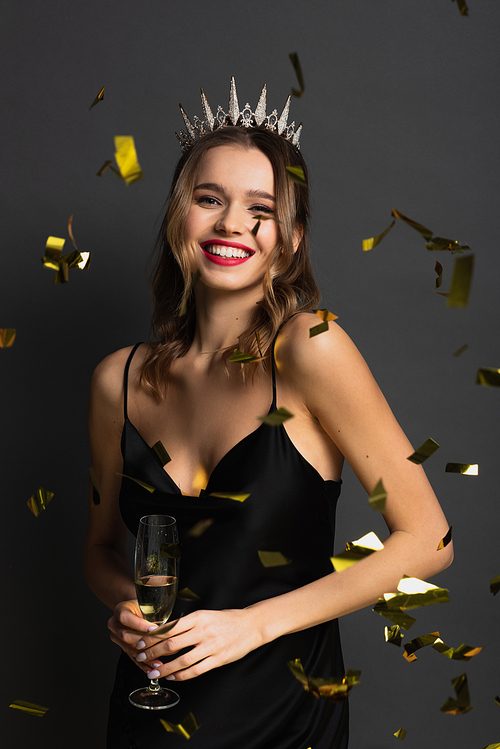 joyful young woman in black slip dress and tiara holding glass of champagne near confetti on grey