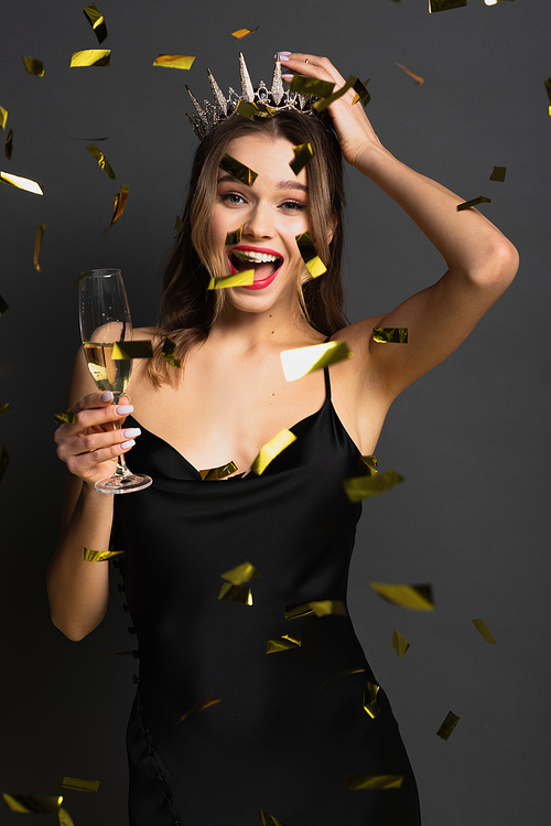 joyful young woman in black slip dress and tiara holding glass of champagne near confetti on grey