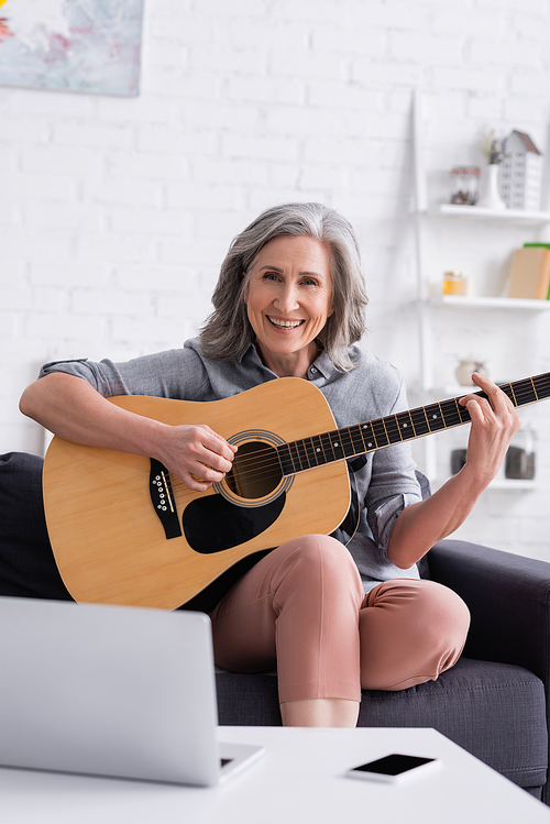 happy mature woman learning to play acoustic guitar near laptop and smartphone with blank screen