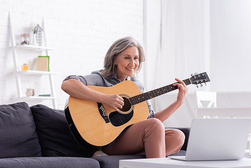 happy middle aged woman learning to play acoustic guitar near laptop on coffee table