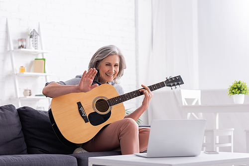 middle aged woman with grey hair holding acoustic guitar while waving hand during video call on laptop