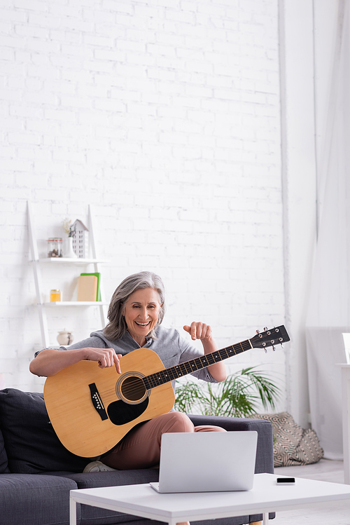 middle aged woman with grey hair pointing at acoustic guitar during video call on laptop