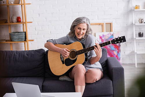 amazed middle aged woman with grey hair learning to play acoustic guitar near laptop