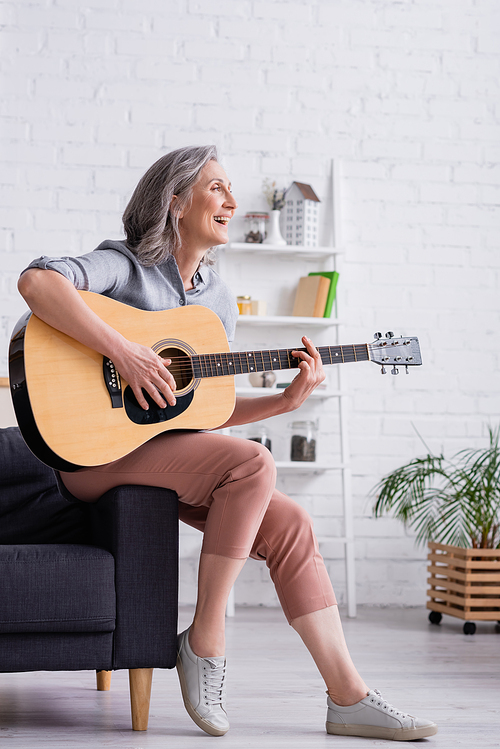 cheerful mature woman with grey hair playing acoustic guitar while sitting in living room