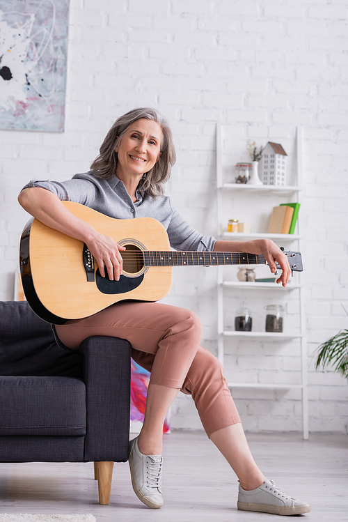 smiling mature woman with grey hair sitting with acoustic guitar in living room