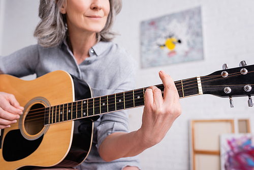 partial view of smiling middle aged woman playing acoustic guitar in living room
