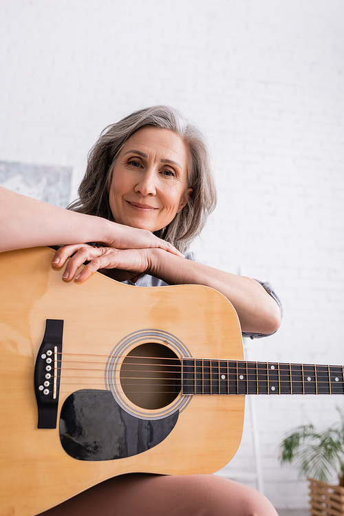 pleased mature woman with grey hair holding acoustic guitar in living room