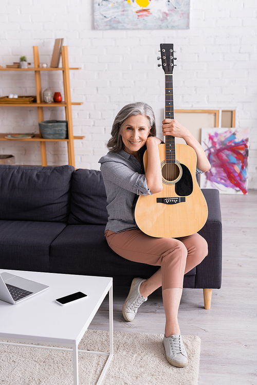 mature woman with grey hair holding acoustic guitar while sitting on couch near gadgets in living room