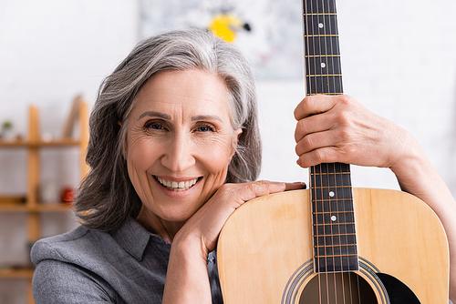 happy mature woman with grey hair holding acoustic guitar in living room