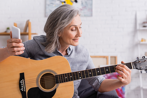 mature woman with grey hair holding smartphone while learning to play acoustic guitar