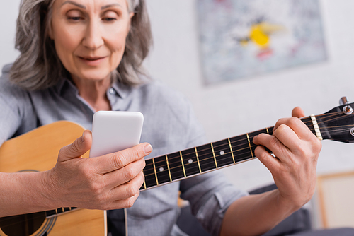 blurred mature woman with grey hair holding smartphone while learning to play acoustic guitar