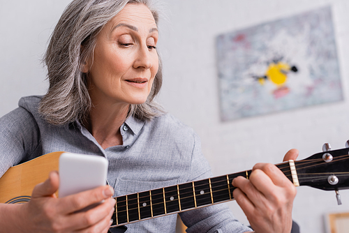 mature woman with grey hair holding blurred smartphone while learning to play acoustic guitar