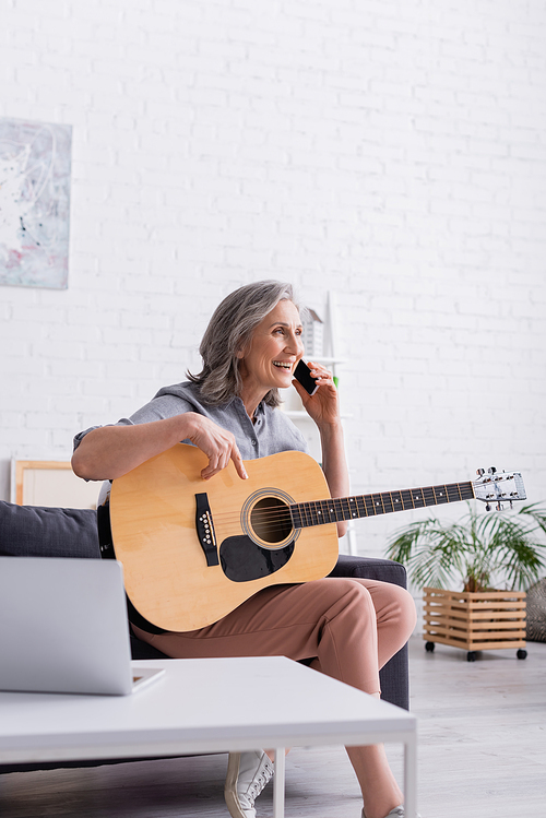 smiling mature woman with grey hair talking on smartphone while holding acoustic guitar