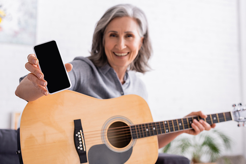blurred and smiling mature woman with grey hair holding smartphone with blank screen and acoustic guitar