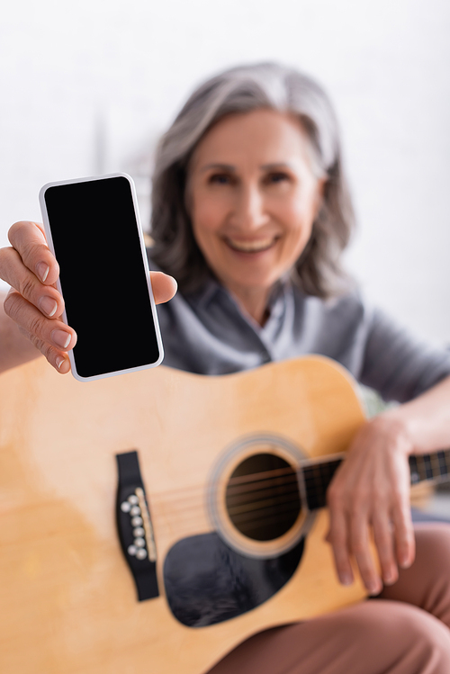 blurred and happy mature woman with grey hair holding smartphone with blank screen and acoustic guitar
