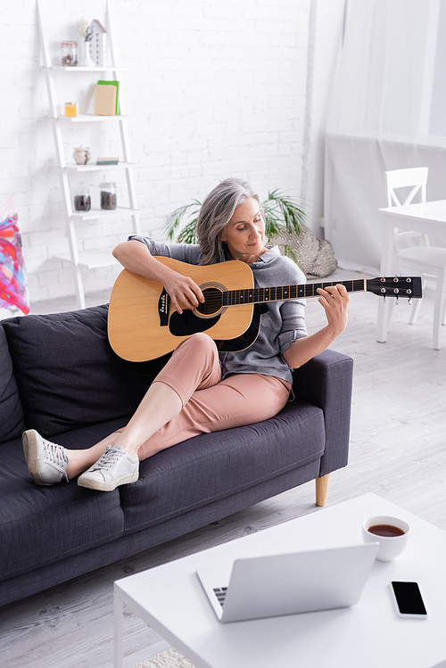 middle aged woman playing acoustic guitar near gadgets and cup of tea on coffee table