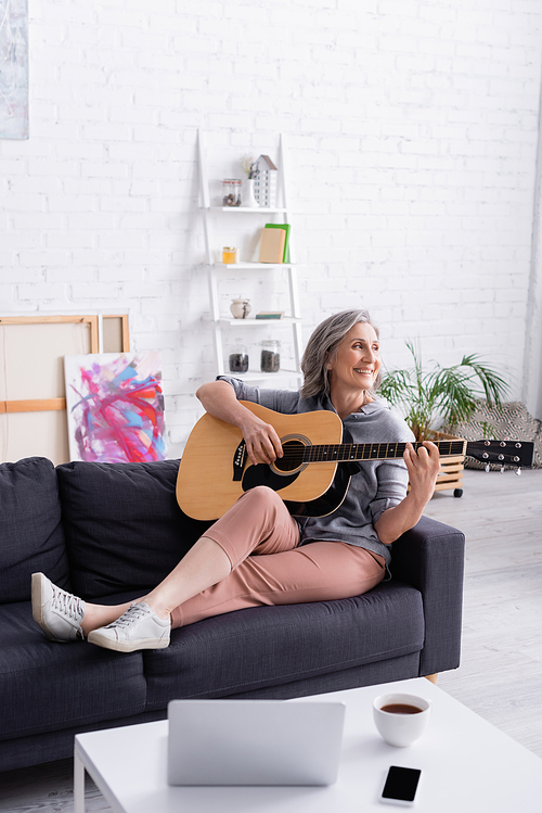 happy middle aged woman playing acoustic guitar near gadgets and cup of tea on coffee table