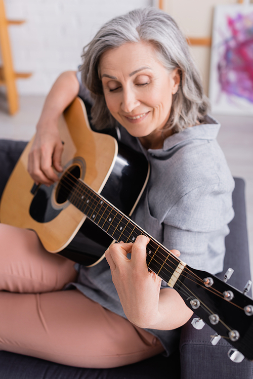 positive mature woman with grey hair playing acoustic guitar in living room