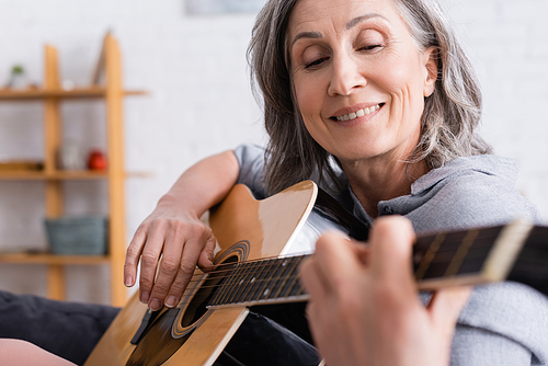 positive mature woman with grey hair playing acoustic guitar on blurred foreground