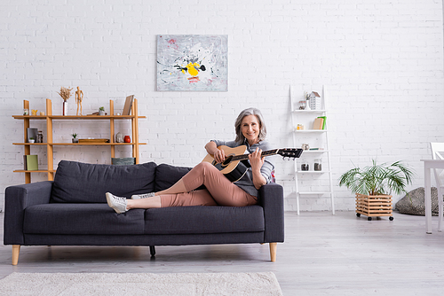 happy mature woman with grey hair sitting on couch and playing acoustic guitar in modern living room