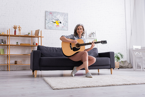 cheerful mature woman with grey hair sitting on couch and playing acoustic guitar in living room
