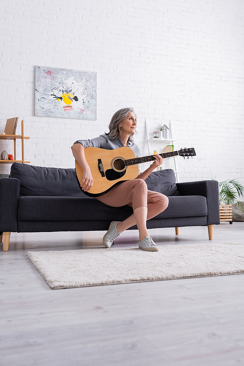 dreamy mature woman with grey hair sitting on couch with acoustic guitar