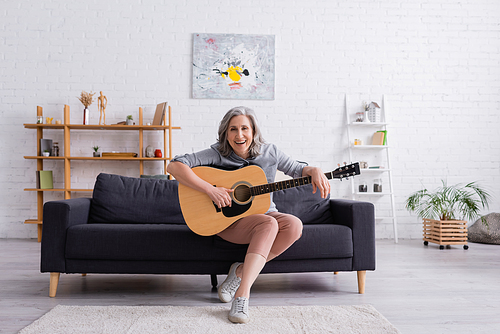 cheerful mature woman with grey hair sitting on couch with acoustic guitar