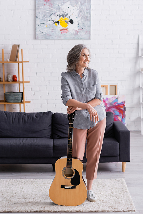 pleased mature woman with grey hair standing with acoustic guitar near couch in living room