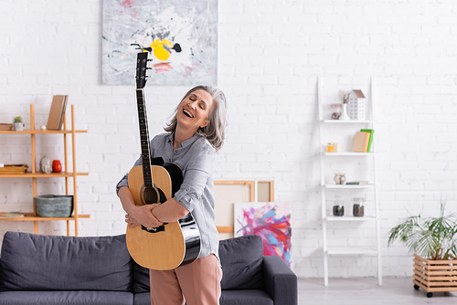 joyful mature woman with grey hair standing with acoustic guitar near couch in living room